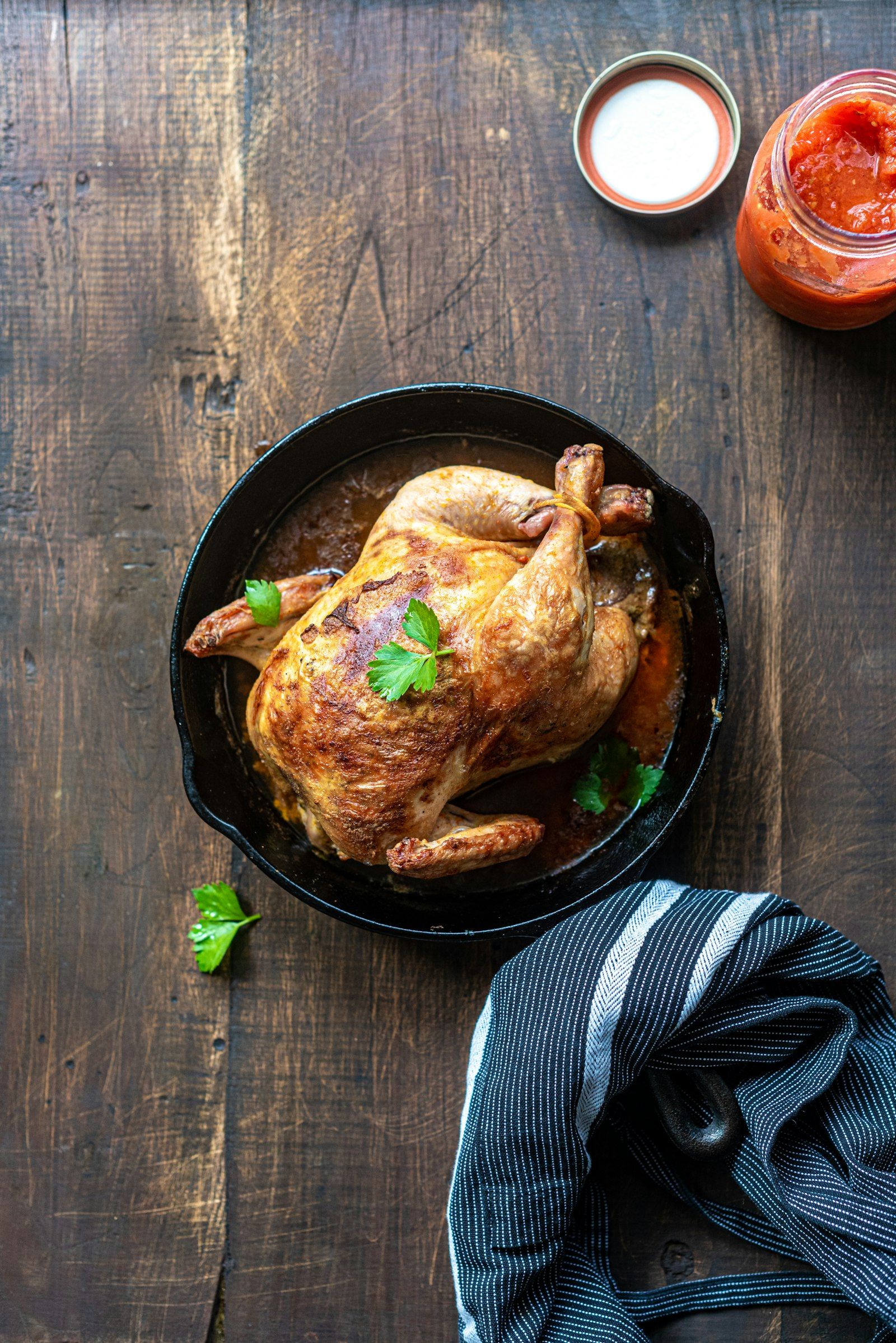 A whole roast chicken with crisp golden-brown skin in a cast iron pan, surrounded by halved potatoes and lemon wedges.